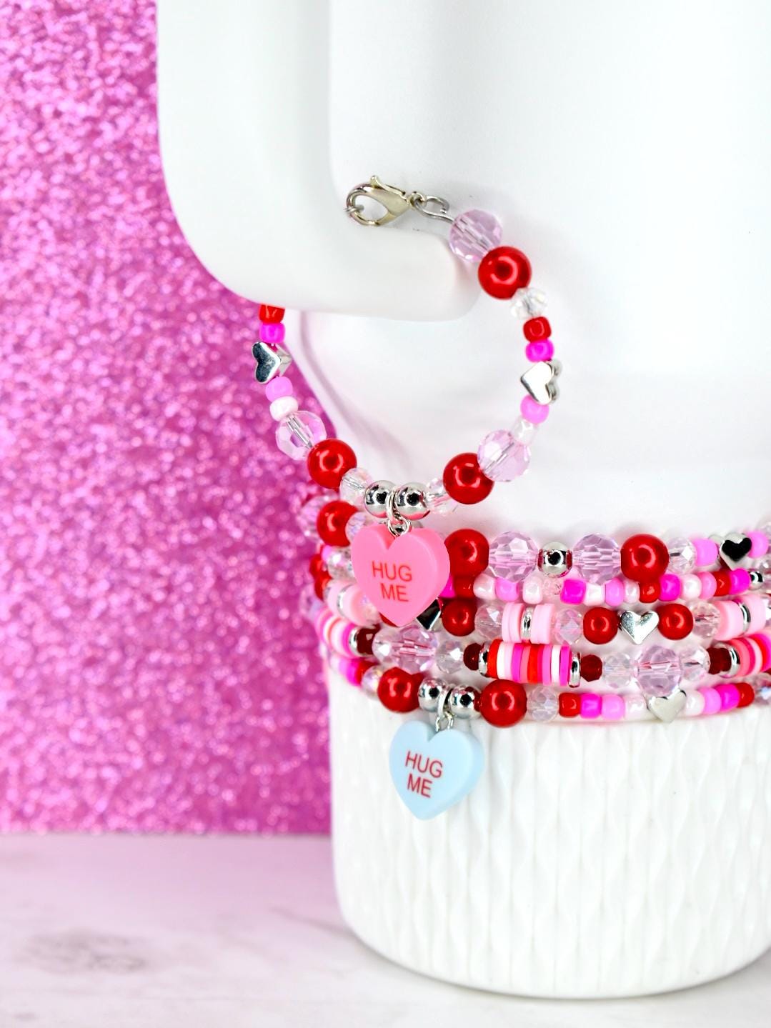 A stack of beaded bracelets in shades of red, pink, and clear. Some bracelets feature heart-shaped charms with the text "HUG ME". The bracelets are displayed on a white surface against a pink glitter background.