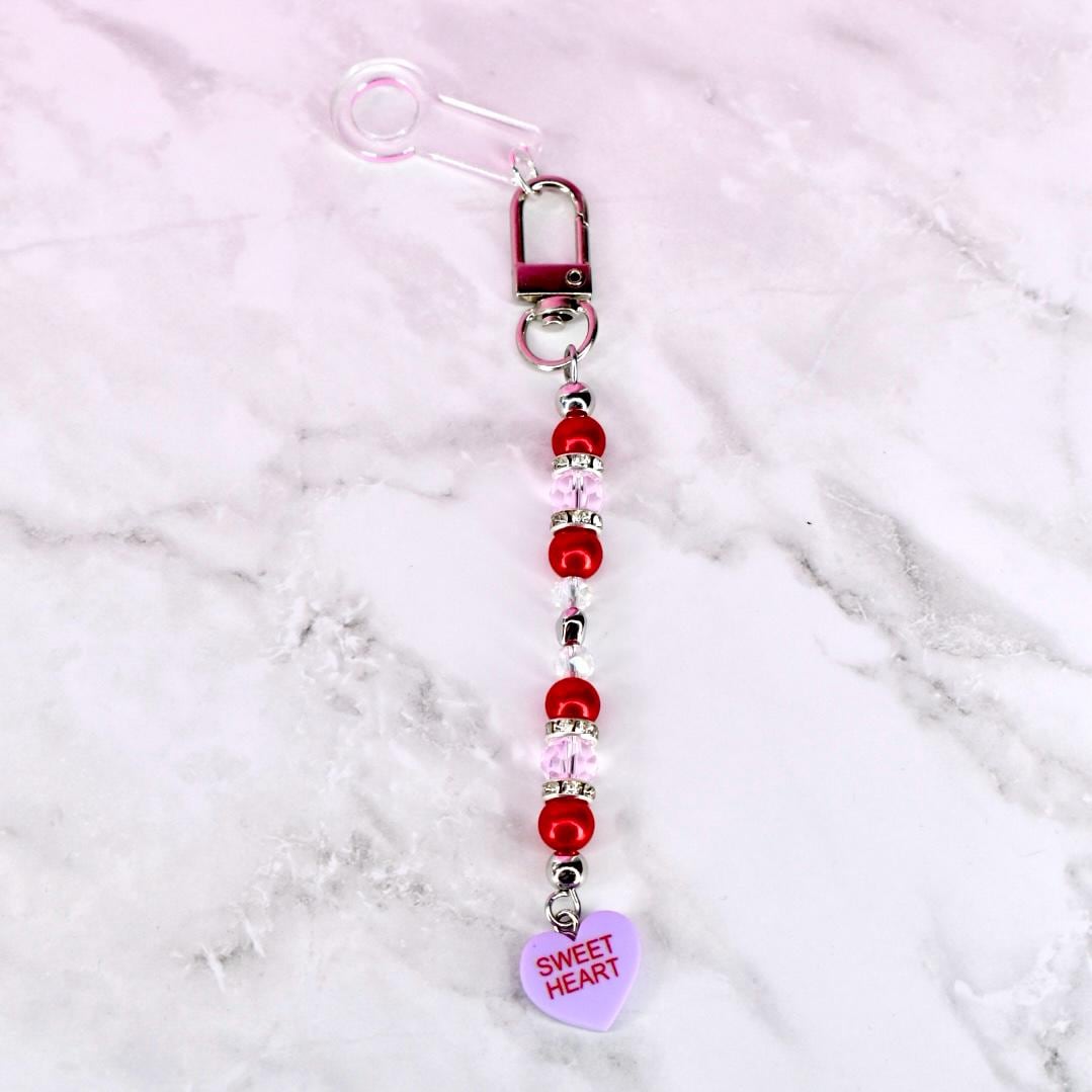 A beaded charm with a clear safety pin, a silver clasp, and a string of red and pink beads. A small, light purple heart-shaped charm at the bottom reads "Sweet Heart".
