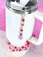 A white tumbler with a silver rim and handle, adorned with a beaded charm. The charm features red, pink, and clear beads, and a purple heart-shaped pendant that reads "Sweet Heart". A matching beaded bracelet is also visible.