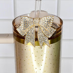 A gold-colored tumbler with a handle and a clear straw. The tumbler is adorned with a gold bow-shaped charm encrusted with sparkling rhinestones. The tumbler has a gold dotted pattern.