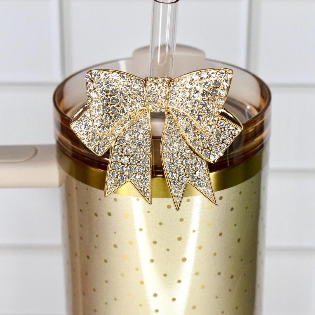 A gold-colored tumbler with a handle and a clear straw. The tumbler is adorned with a gold bow-shaped charm encrusted with sparkling rhinestones. The tumbler has a gold dotted pattern.