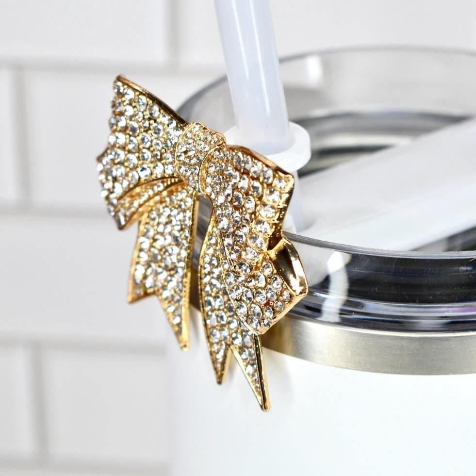 A gold-colored bow-shaped accessory, encrusted with clear rhinestones, is attached to a white tumbler with a clear straw. The bow adds a touch of glamour to the drinkware. The tumbler has a stainless steel rim.