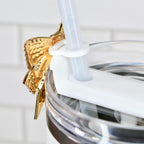 Close-up of a white tumbler with a clear lid and a white straw. A gold-colored, textured star-shaped charm with rhinestones is attached to the lid. The tumbler is white with a silver band.