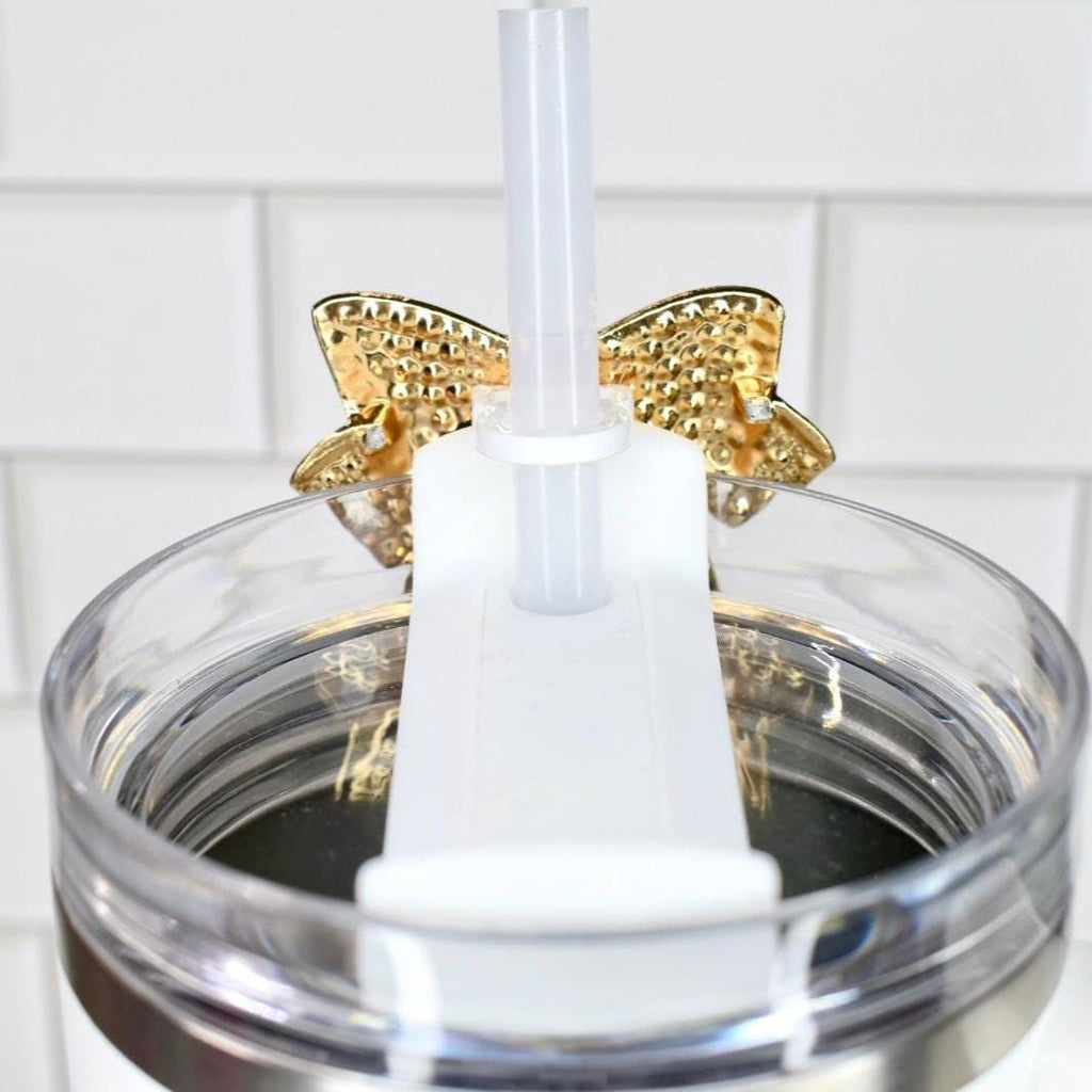 Close-up of a clear tumbler with a gold bow accent. A white straw and a white plastic piece are inserted into the tumbler. The tumbler is set against a white tile background.