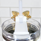 Close-up of a clear tumbler with a gold bow accent. A white straw and a white plastic piece are inserted into the tumbler. The tumbler is set against a white tile background.