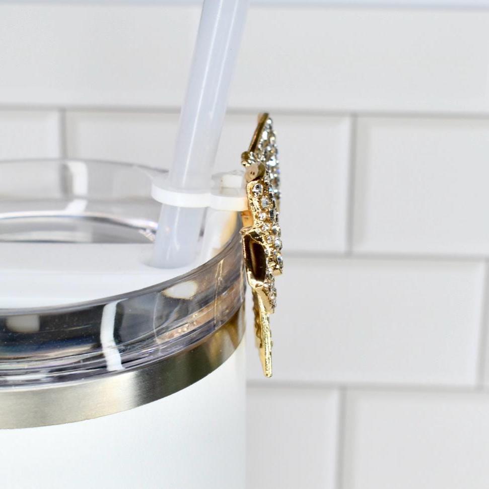 A white tumbler with a gold rim and a decorative gold butterfly charm. The charm is adorned with clear rhinestones. A white straw is inserted into the tumbler.