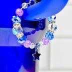 A beaded bracelet with blue, purple, and clear beads. The bracelet has a star charm with iridescent coloring. The bracelet is hanging on a blue object.
