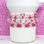 A stack of five beaded bracelets in shades of pink and clear. The bracelets feature a variety of beads, including clear crystal, pink round beads, and hot pink flat beads. The bracelets are stacked on a white ceramic mug.
