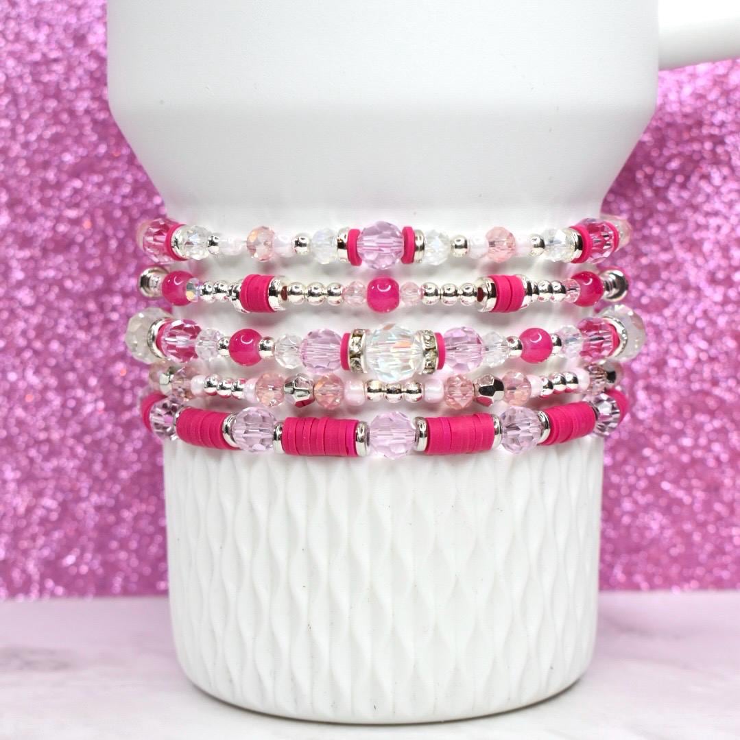 A stack of five beaded bracelets in shades of pink and clear. The bracelets feature a variety of beads, including clear crystal, pink round beads, and hot pink flat beads. The bracelets are stacked on a white ceramic mug.