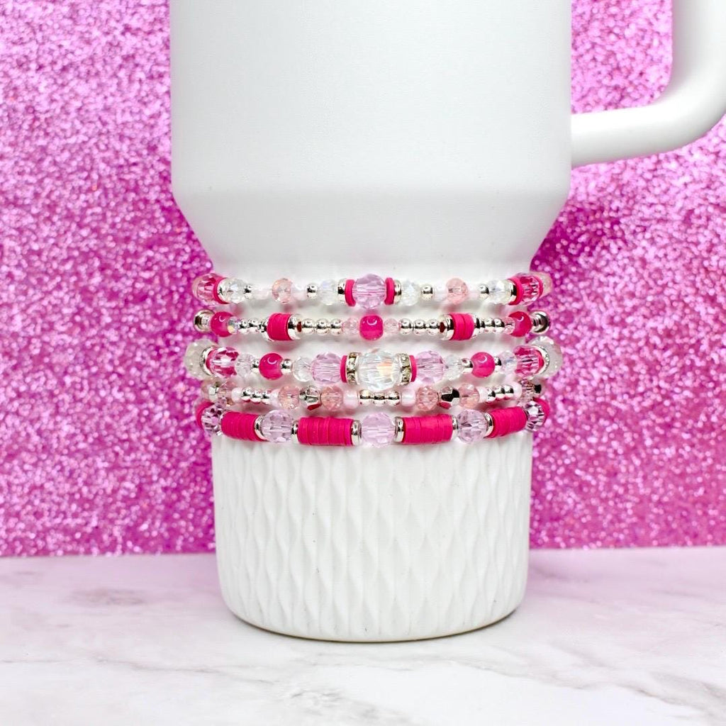 A stack of five beaded bracelets in shades of pink, clear, and silver. The bracelets are displayed on a white ceramic mug against a sparkly pink background. The beads include clear, pink, and silver accents.