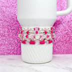 A stack of five beaded bracelets in shades of pink, clear, and silver. The bracelets are displayed on a white ceramic mug against a sparkly pink background. The beads include clear, pink, and silver accents.