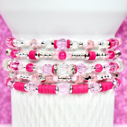 Stack of five beaded bracelets in shades of pink and clear. The bracelets feature a variety of beads, including clear crystal, pink round beads, and silver accents. The bracelets are stacked on a white cylindrical surface.
