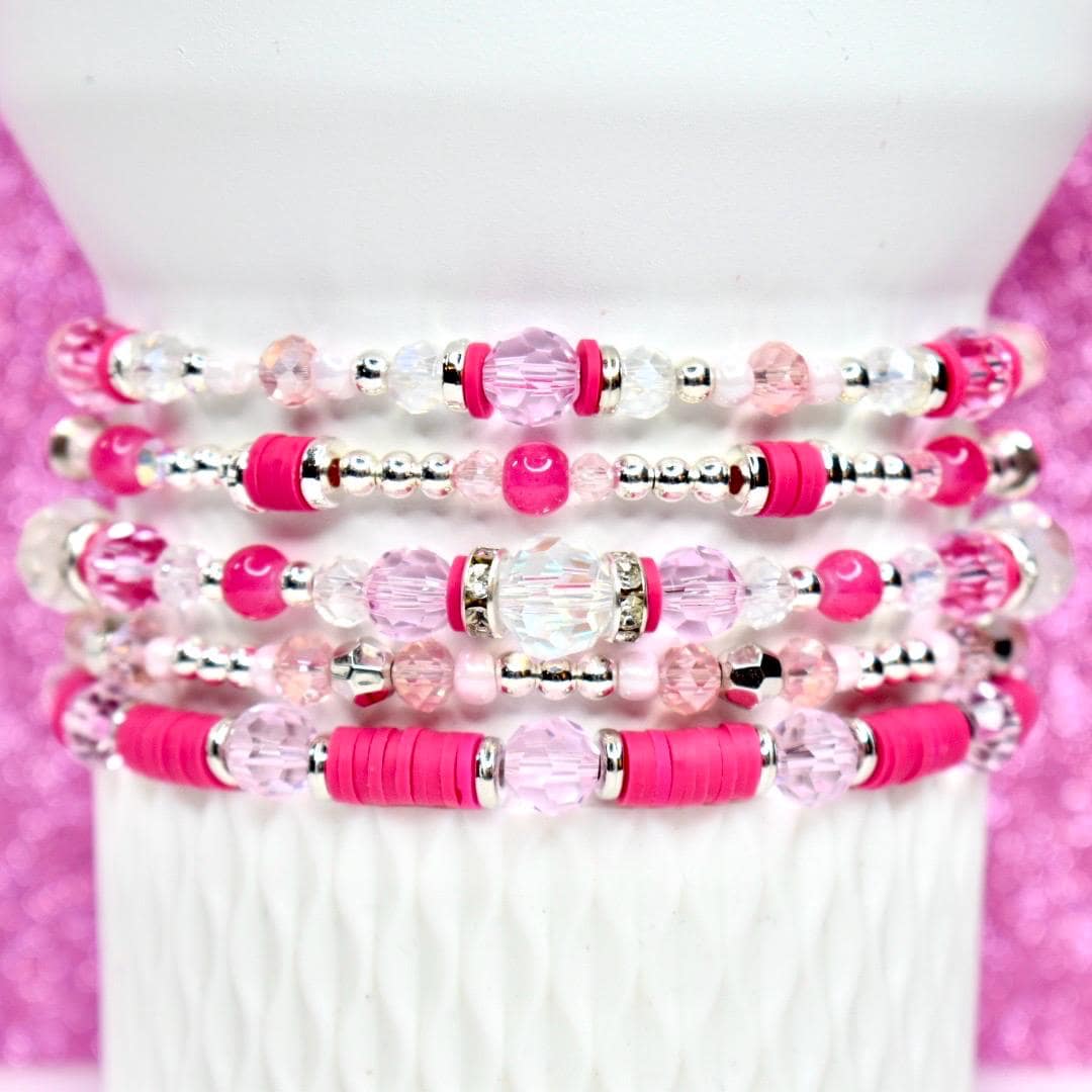 Stack of five beaded bracelets in shades of pink and clear. The bracelets feature a variety of beads, including clear crystal, pink round beads, and silver accents. The bracelets are stacked on a white cylindrical surface.