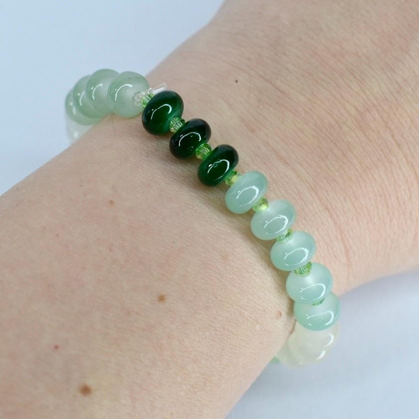 A beaded bracelet featuring a gradient of green hues. The bracelet transitions from light green to dark green beads, strung together with a clear cord. The beads have a smooth, rounded shape.