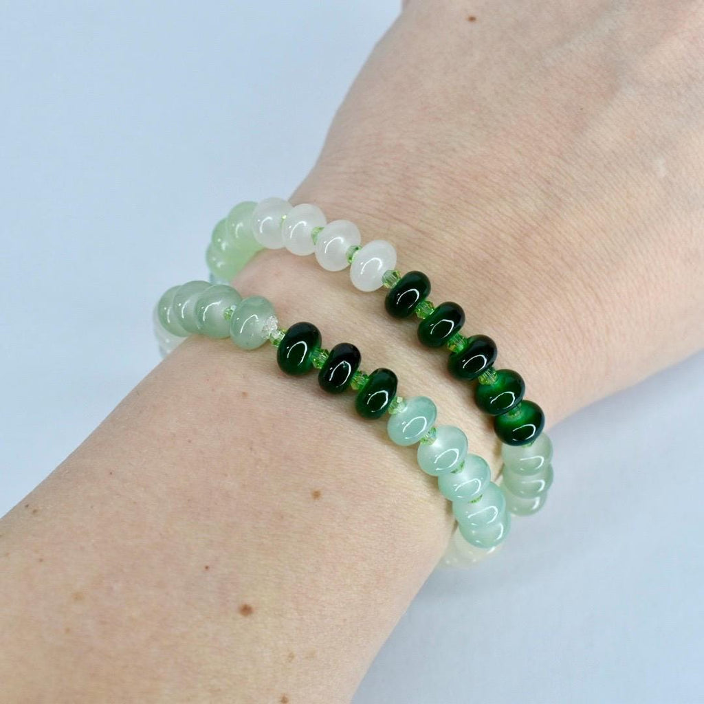 Two beaded bracelets. One bracelet features light green and white beads, while the other has light green and dark green beads. The bracelets are worn on a wrist, showcasing the jewelry's design and color variations.