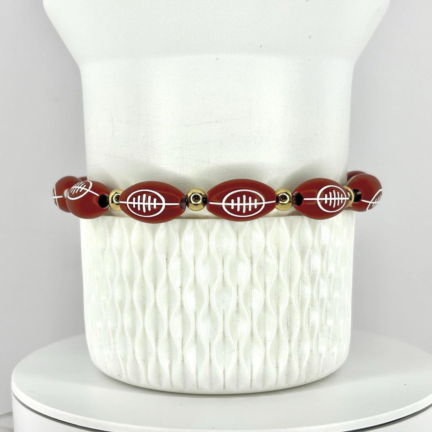 A football-themed bracelet. The bracelet features oval-shaped beads with white football designs, interspersed with gold-colored beads. The bracelet is displayed on a white, textured surface.