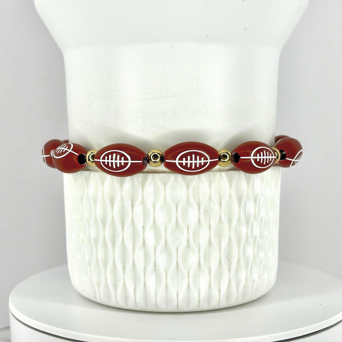 A football-themed bracelet. The bracelet features oval-shaped beads with white football designs, interspersed with gold-colored beads. The bracelet is displayed on a white, textured surface.