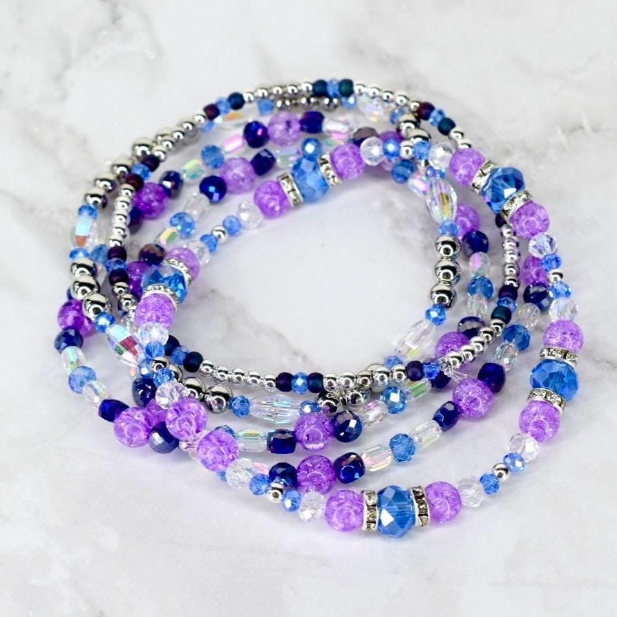 A collection of beaded bracelets in shades of purple, blue, and silver. The bracelets feature various bead sizes and textures, including faceted and round beads. The arrangement is layered, creating a visually appealing stack of jewelry.