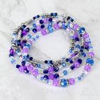A collection of beaded bracelets in shades of purple, blue, and silver. The bracelets feature various bead sizes and textures, including faceted and round beads. The arrangement is layered, creating a visually appealing stack of jewelry.