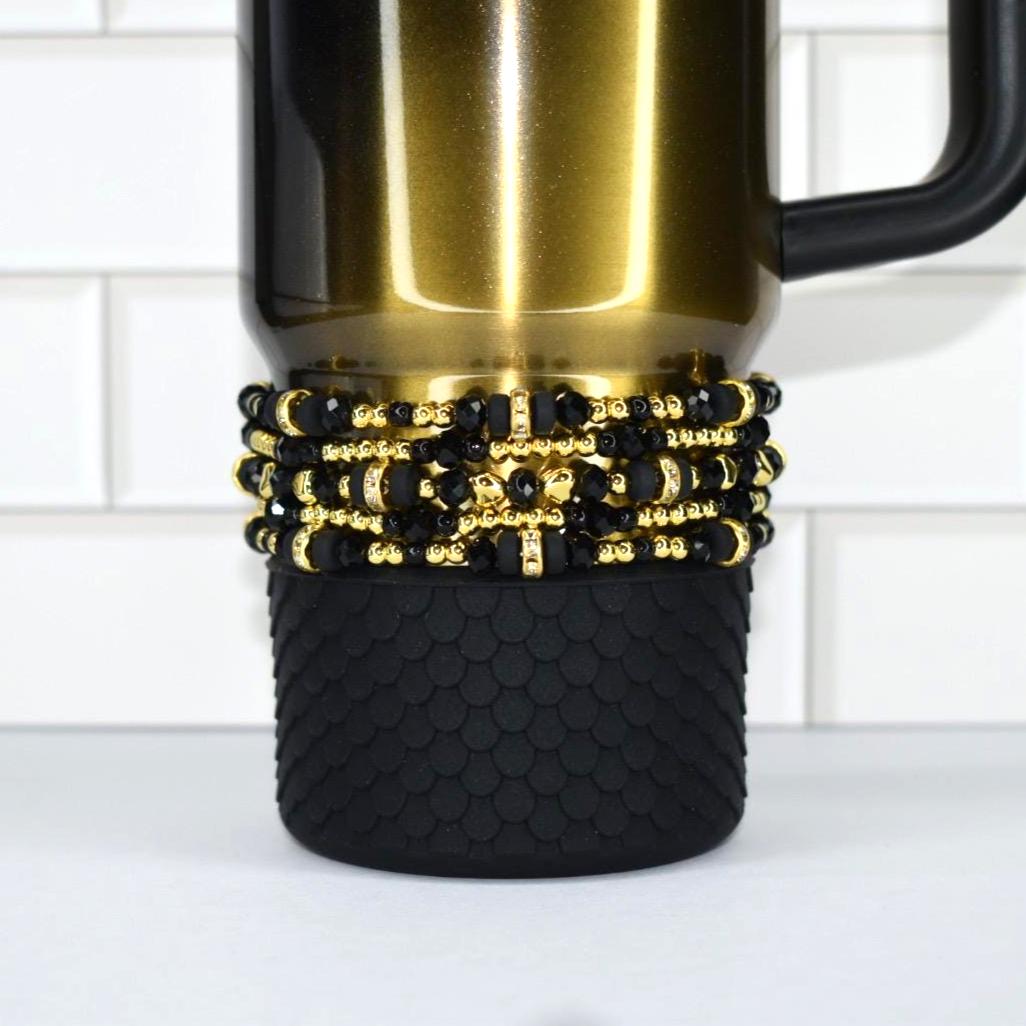 A gold-colored travel mug with a black handle. The mug is adorned with a black and gold beaded wrap and a black textured base. The mug is set against a white tile background.
