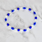 A beaded bracelet featuring a combination of clear, faceted beads, vibrant blue beads, and cylindrical blue accents. The bracelet is strung together, creating a circular shape. The beads are arranged in a repeating pattern.