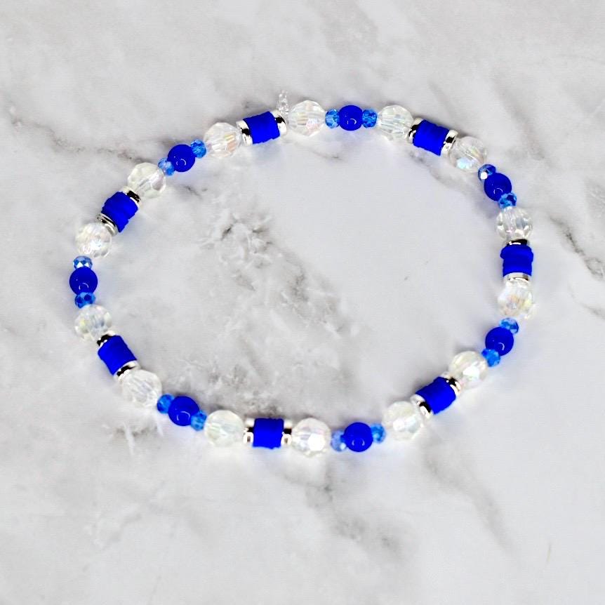 A beaded bracelet featuring a combination of clear, faceted beads, vibrant blue beads, and cylindrical blue accents. The bracelet is strung together, creating a circular shape. The beads are arranged in a repeating pattern.