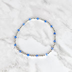A delicate bracelet featuring small, silver-colored beads interspersed with vibrant blue crystal beads. The bracelet is circular and rests on a white and gray marble surface. The design is simple and elegant.