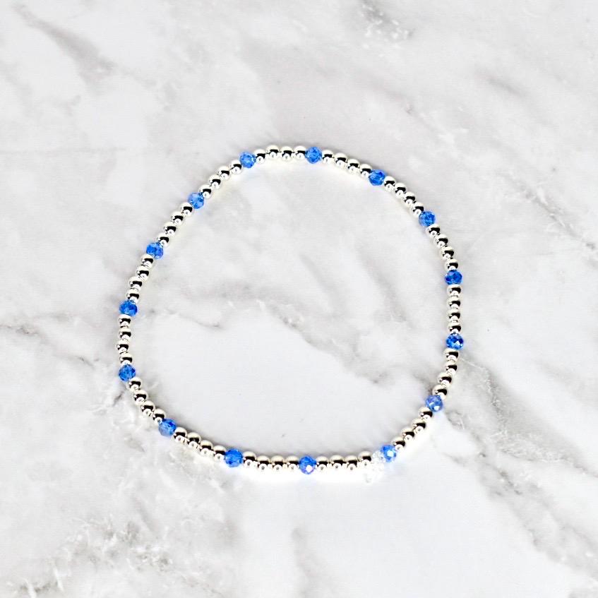 A delicate bracelet featuring small, silver-colored beads interspersed with vibrant blue crystal beads. The bracelet is circular and rests on a white and gray marble surface. The design is simple and elegant.
