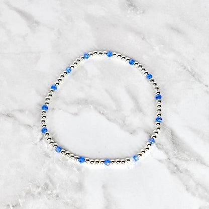 A delicate bracelet featuring small, silver-colored beads interspersed with vibrant blue crystal beads. The bracelet is circular and rests on a white and gray marble surface. The design is simple and elegant.