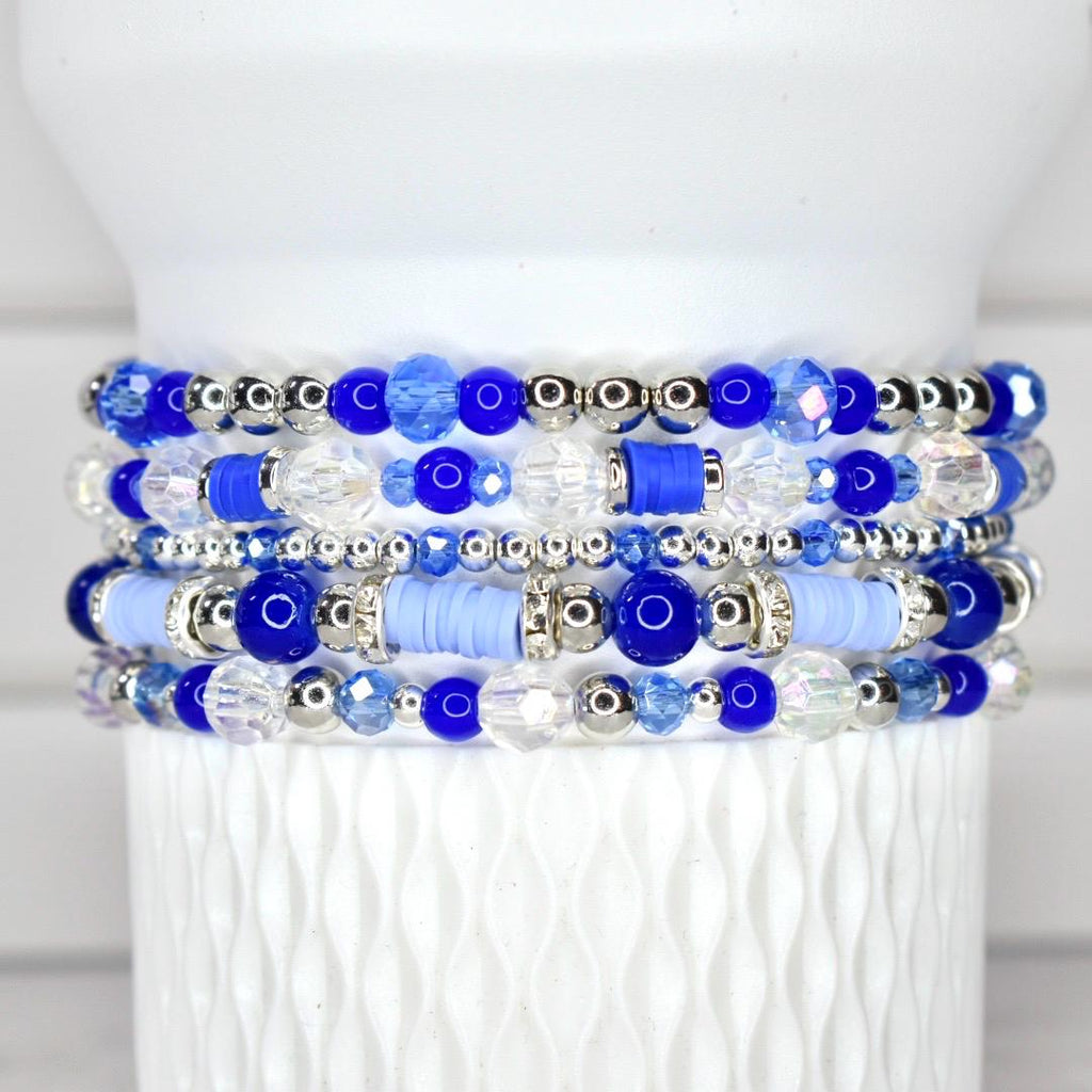 Stack of five beaded bracelets in shades of blue and clear. The bracelets feature a variety of beads, including round, faceted, and disc-shaped beads, with silver-colored accents. The bracelets are displayed on a white, textured surface.