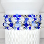 Stack of five beaded bracelets in shades of blue and clear. The bracelets feature a variety of beads, including round, faceted, and disc-shaped beads, with silver-colored accents. The bracelets are displayed on a white, textured surface.