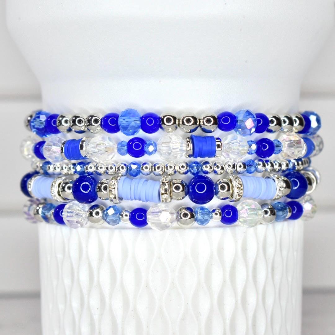 Stack of five beaded bracelets in shades of blue and clear. The bracelets feature a variety of beads, including round, faceted, and disc-shaped beads, with silver-colored accents. The bracelets are displayed on a white, textured surface.