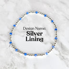 A silver beaded bracelet with blue crystal accents. The bracelet is circular and features the text "Design Name: Silver Lining" in black font. The bracelet is displayed on a white and gray marble surface.