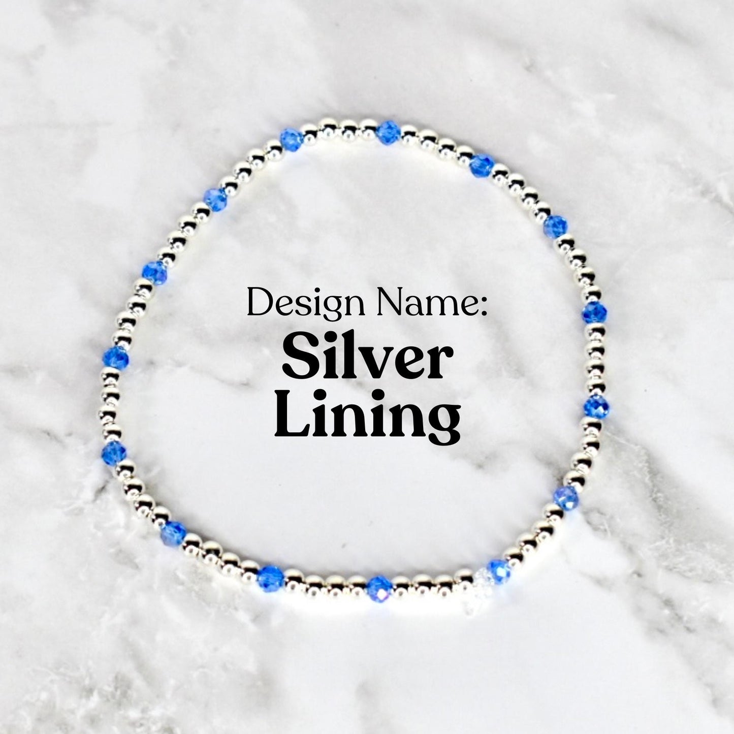 A silver beaded bracelet with blue crystal accents. The bracelet is circular and features the text "Design Name: Silver Lining" in black font. The bracelet is displayed on a white and gray marble surface.