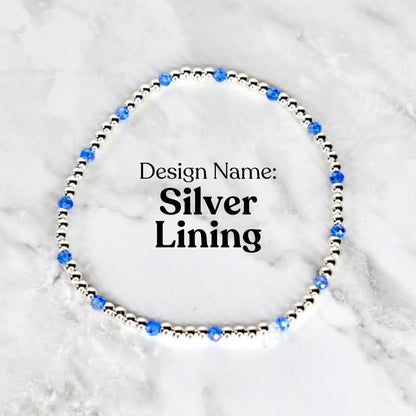 A silver beaded bracelet with blue crystal accents. The bracelet is circular and features the text "Design Name: Silver Lining" in black font. The bracelet is displayed on a white and gray marble surface.