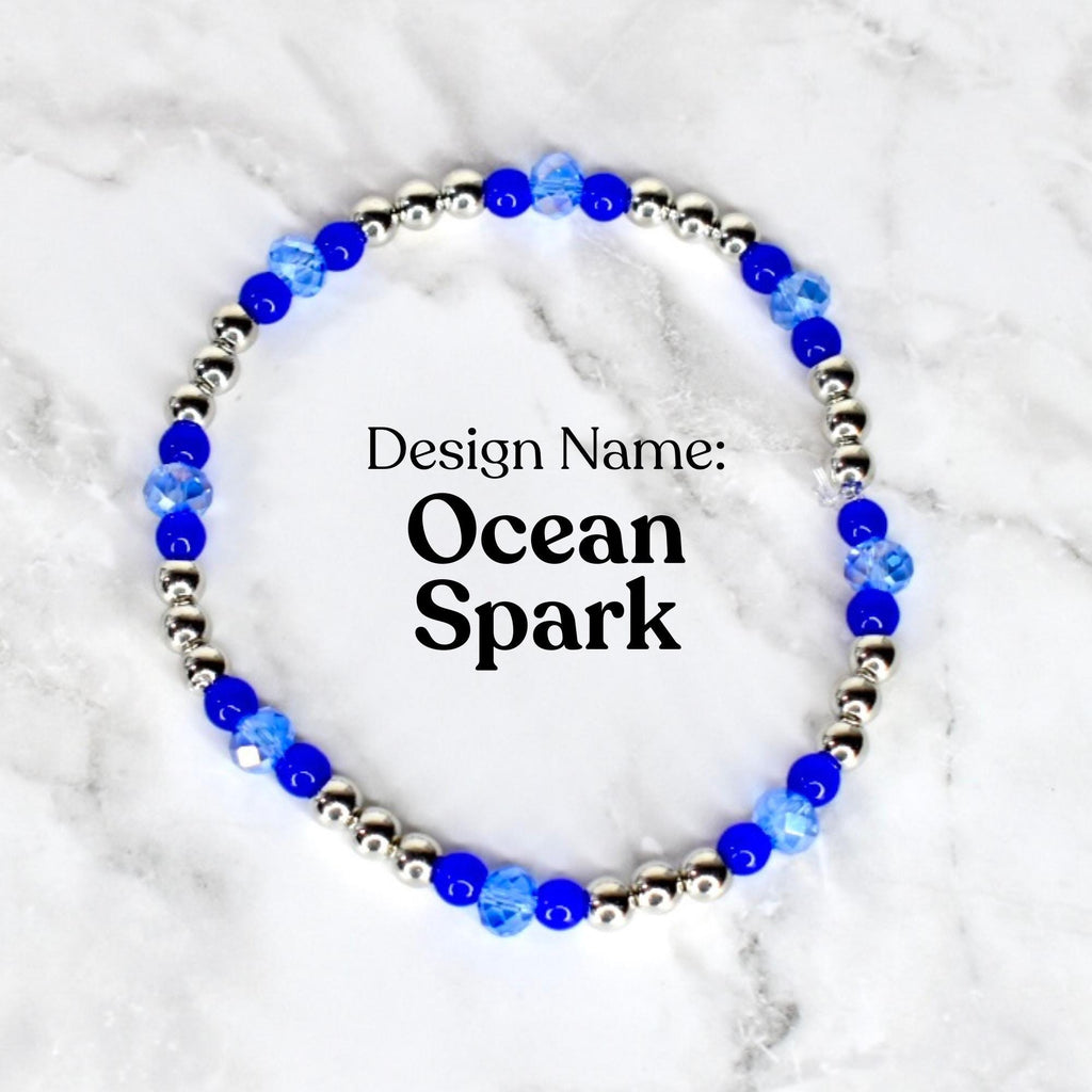 A beaded bracelet with the text "Design Name: Ocean Spark". The bracelet features alternating silver-colored beads and blue faceted beads. The bracelet is displayed on a white and gray marble surface.