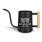 Black watering can with a long, curved spout and a wooden handle. The can features a white floral design and the text "Love grows here Est. 2025". A stylish and functional gardening accessory.