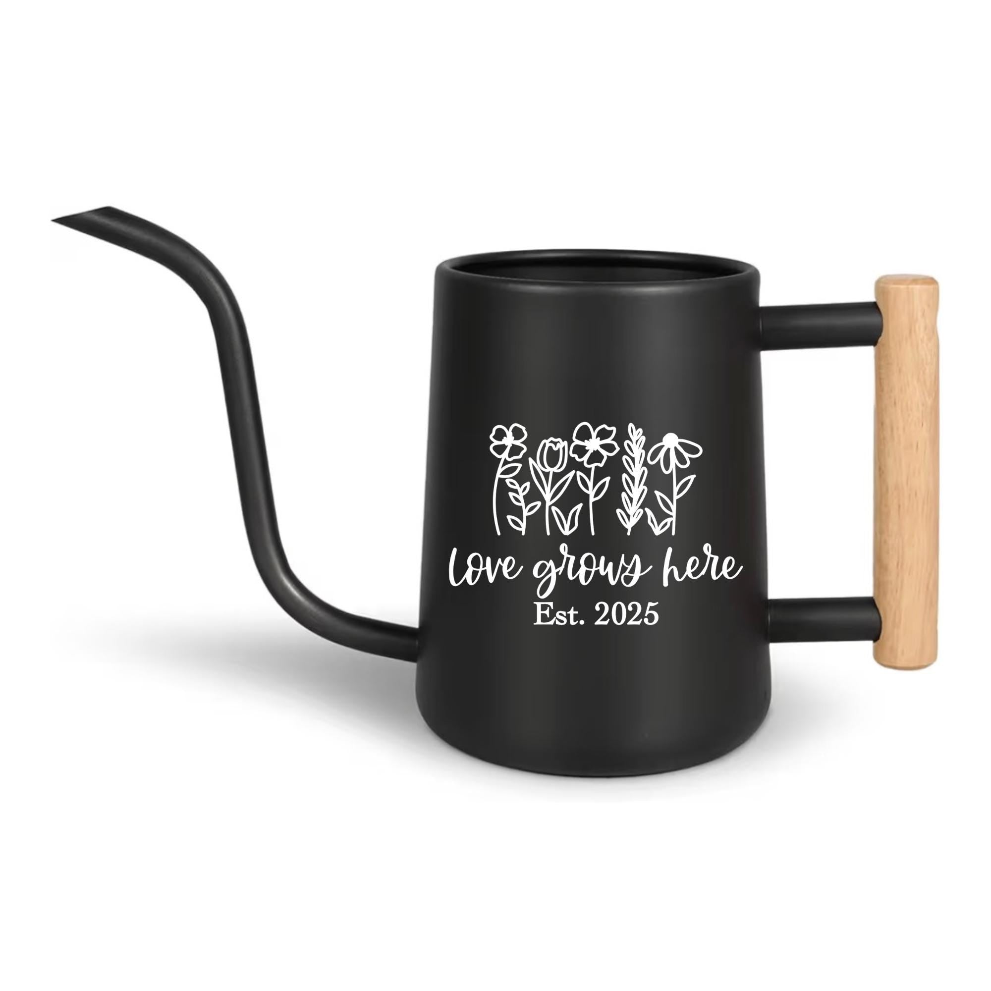 Black watering can with a long, curved spout and a wooden handle. The can features a white floral design and the text "Love grows here Est. 2025". A stylish and functional gardening accessory.