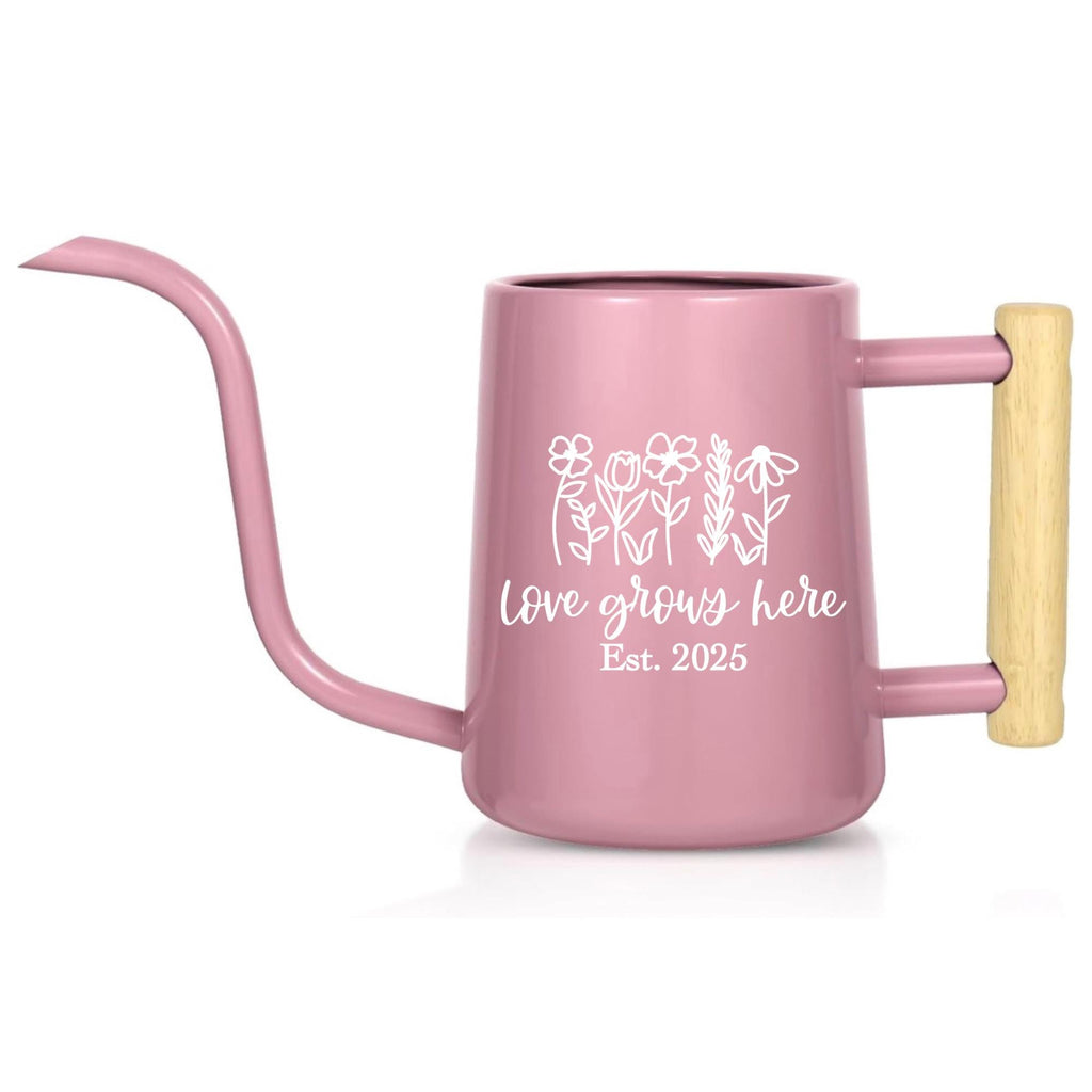 A pink watering can with a long, curved spout and a wooden handle. The can features a white floral design and the text "Love grows here Est. 2025". Ideal for indoor or outdoor gardening.