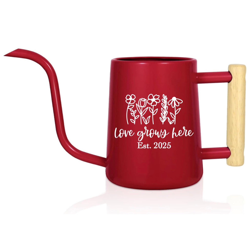 A red watering can with a long, curved spout and a wooden handle. The can features a white floral design and the text "Love grows here Est. 2025".
