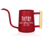 A red watering can with a long, curved spout and a wooden handle. The can features a white floral design and the text "Love grows here Est. 2025".