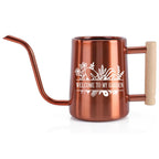 Copper-colored watering can with a long, slender spout and a wooden handle. The can features a white floral design and the text "WELCOME TO MY GARDEN". Ideal for indoor or outdoor gardening.
