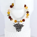 A beaded tumbler handle bracelet featuring a Highland cow charm. The bracelet has a mix of brown and cream-colored beads. The cow charm is silver-toned with detailed features. The bracelet is displayed on a white surface.