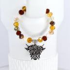 A beaded tumbler handle bracelet featuring a Highland cow charm. The bracelet has a mix of brown and cream-colored beads. The cow charm is silver-toned with detailed features. The bracelet is displayed on a white surface.