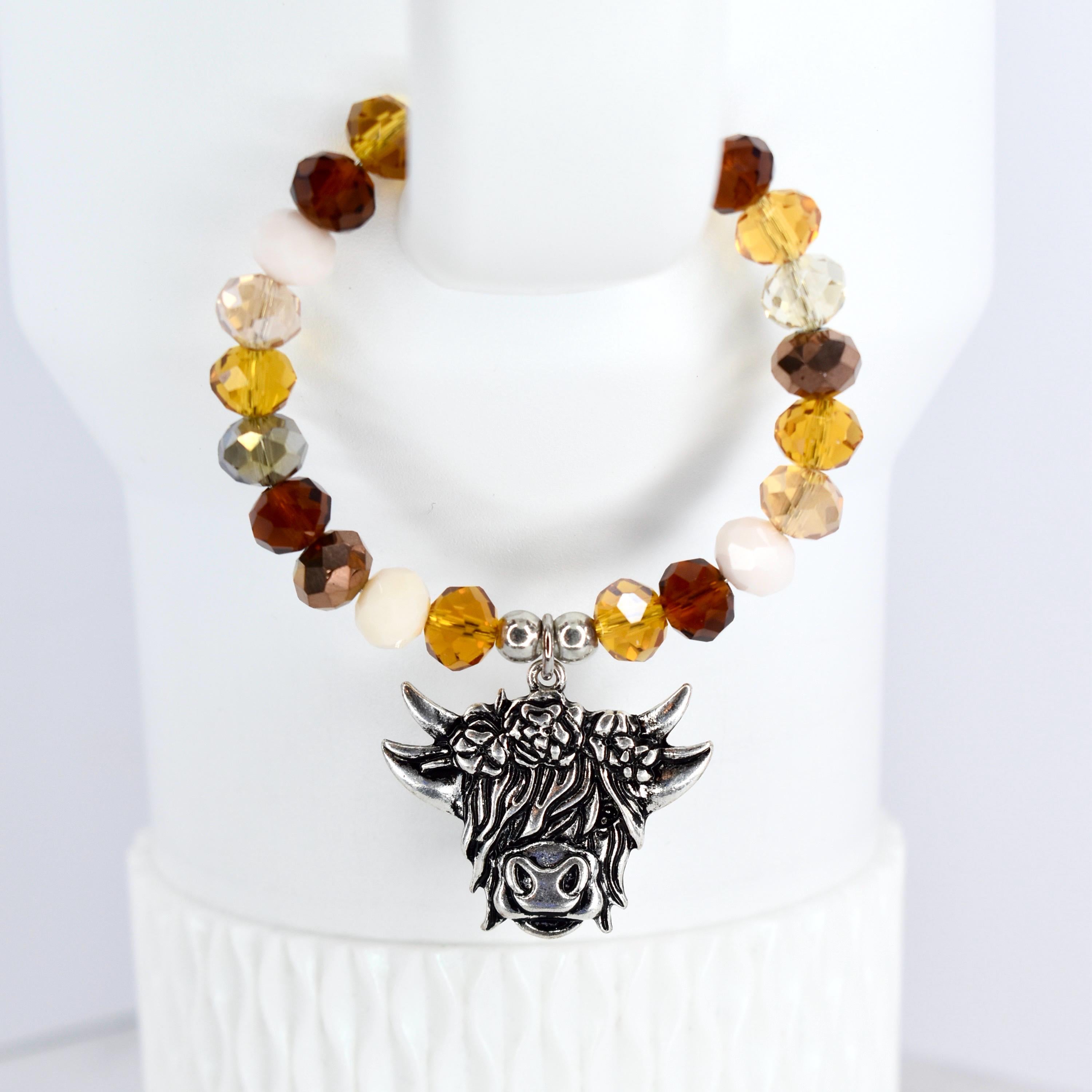A beaded tumbler handle bracelet featuring a Highland cow charm. The bracelet has a mix of brown and cream-colored beads. The cow charm is silver-toned with detailed features. The bracelet is displayed on a white surface.