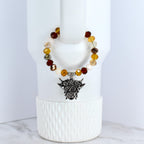 A beaded tumbler bracelet with a silver-colored Highland cow charm. The bracelet features a variety of colorful beads in shades of brown and cream. The charm is detailed with the cow's face and horns.