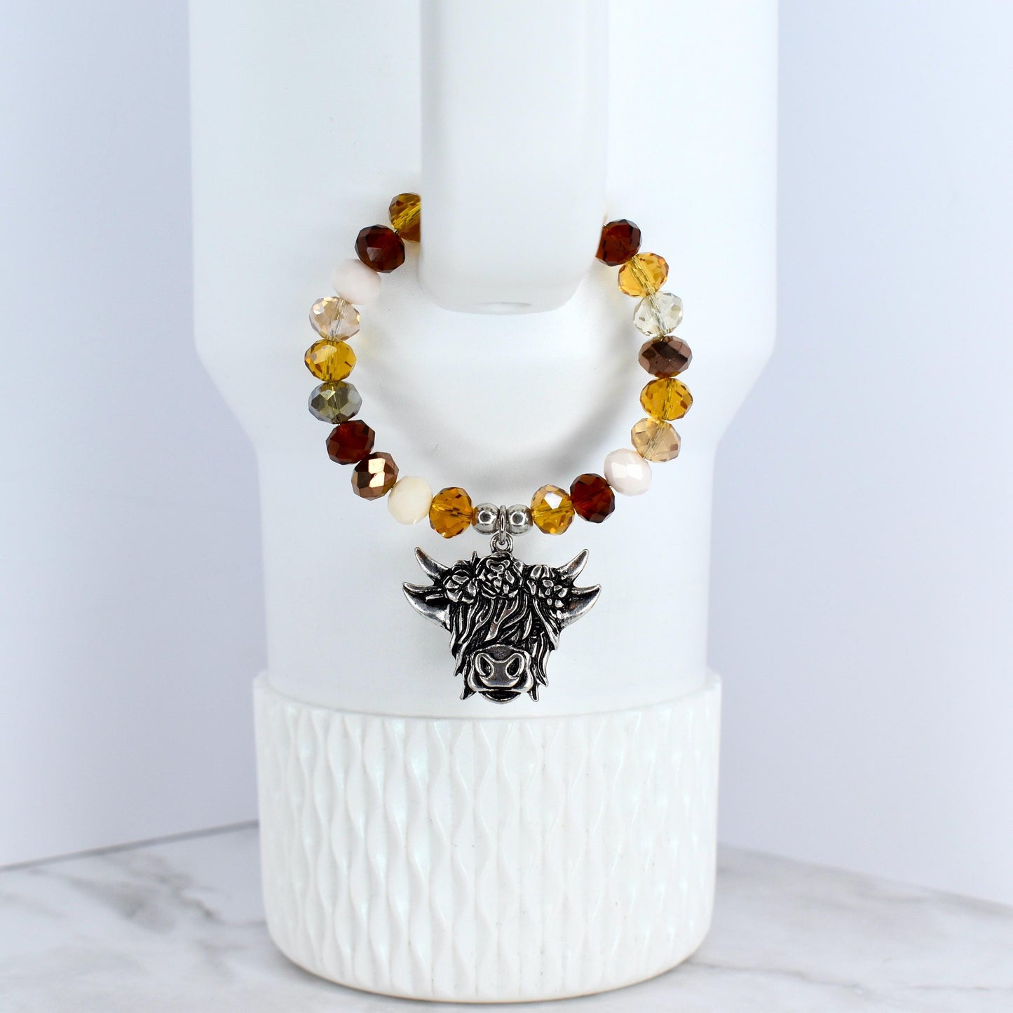 A beaded tumbler bracelet with a silver-colored Highland cow charm. The bracelet features a variety of colorful beads in shades of brown and cream. The charm is detailed with the cow's face and horns.