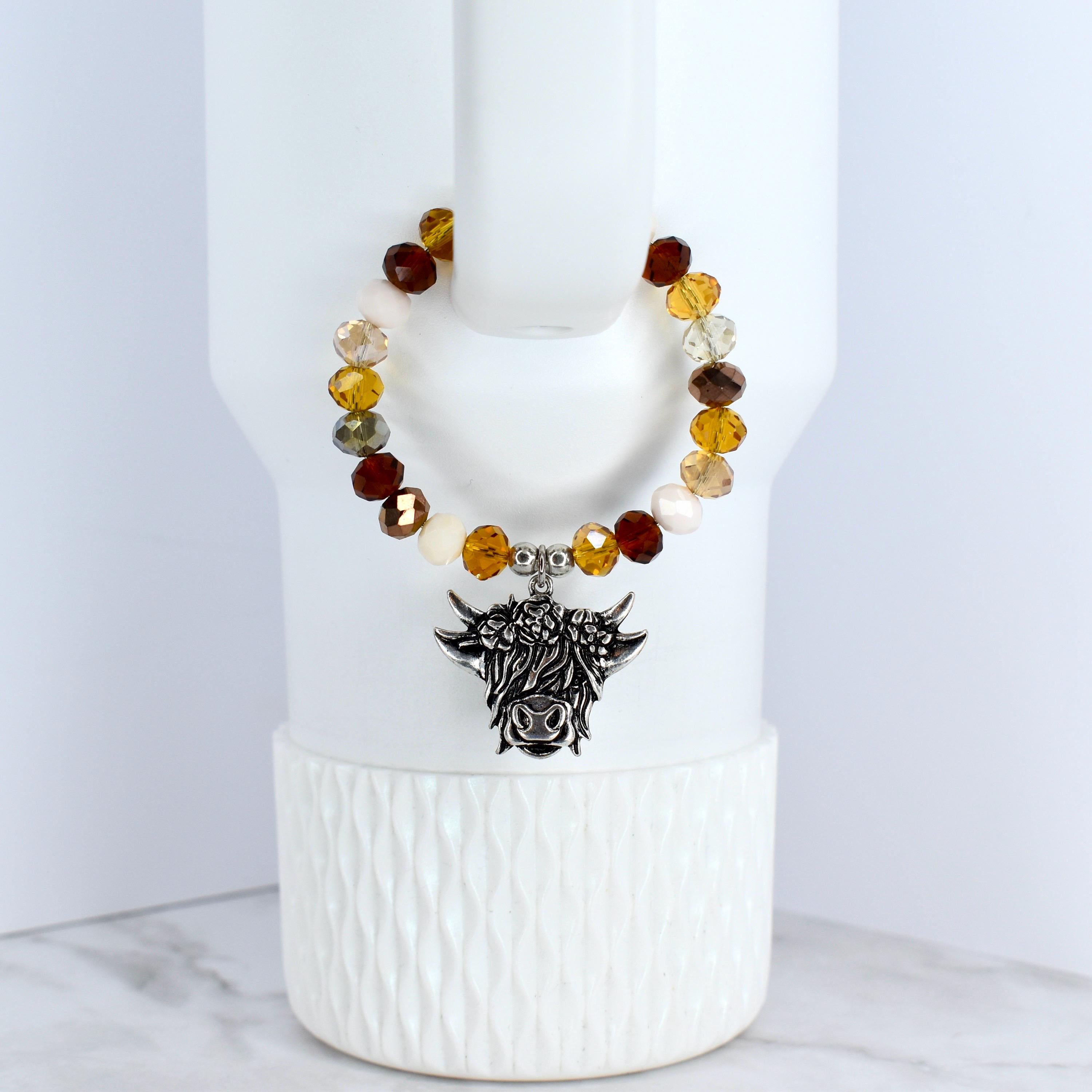 A beaded tumbler bracelet with a silver-colored Highland cow charm. The bracelet features a variety of colorful beads in shades of brown and cream. The charm is detailed with the cow's face and horns.