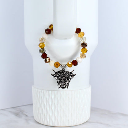 A beaded tumbler bracelet with a silver-colored Highland cow charm. The bracelet features a variety of colorful beads in shades of brown and cream. The charm is detailed with the cow's face and horns.