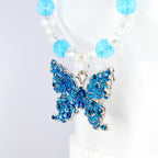 A blue butterfly pendant tumbler handle charm with intricate detailing and silver-colored trim. The butterfly is adorned with small blue crystals. The handle charm features a strand of clear and light blue beads.